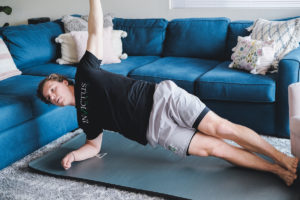 A male athlete wearing an Invictus shirt is doing side planks in his living room.