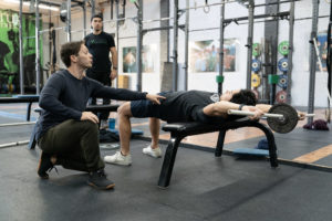 A coach works with an athlete on a mobility drill using a bench, PVC, and change plate while another athlete watches.