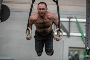 Shirtless male athlete coming down from the top of a ring muscle-up.